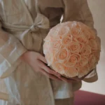 Person in beige satin robe holding bouquet of soft pink roses against blurred background