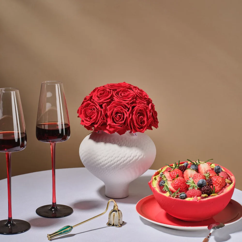 Minimalist elegant still life with white textured vase holding vibrant red rose bouquet beside wine glasses and strawberries