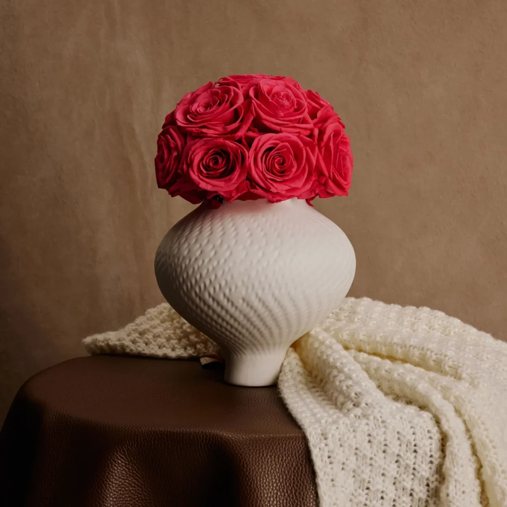 White ceramic textured vase with vibrant red roses on brown leather tablecloth creating rustic atmosphere