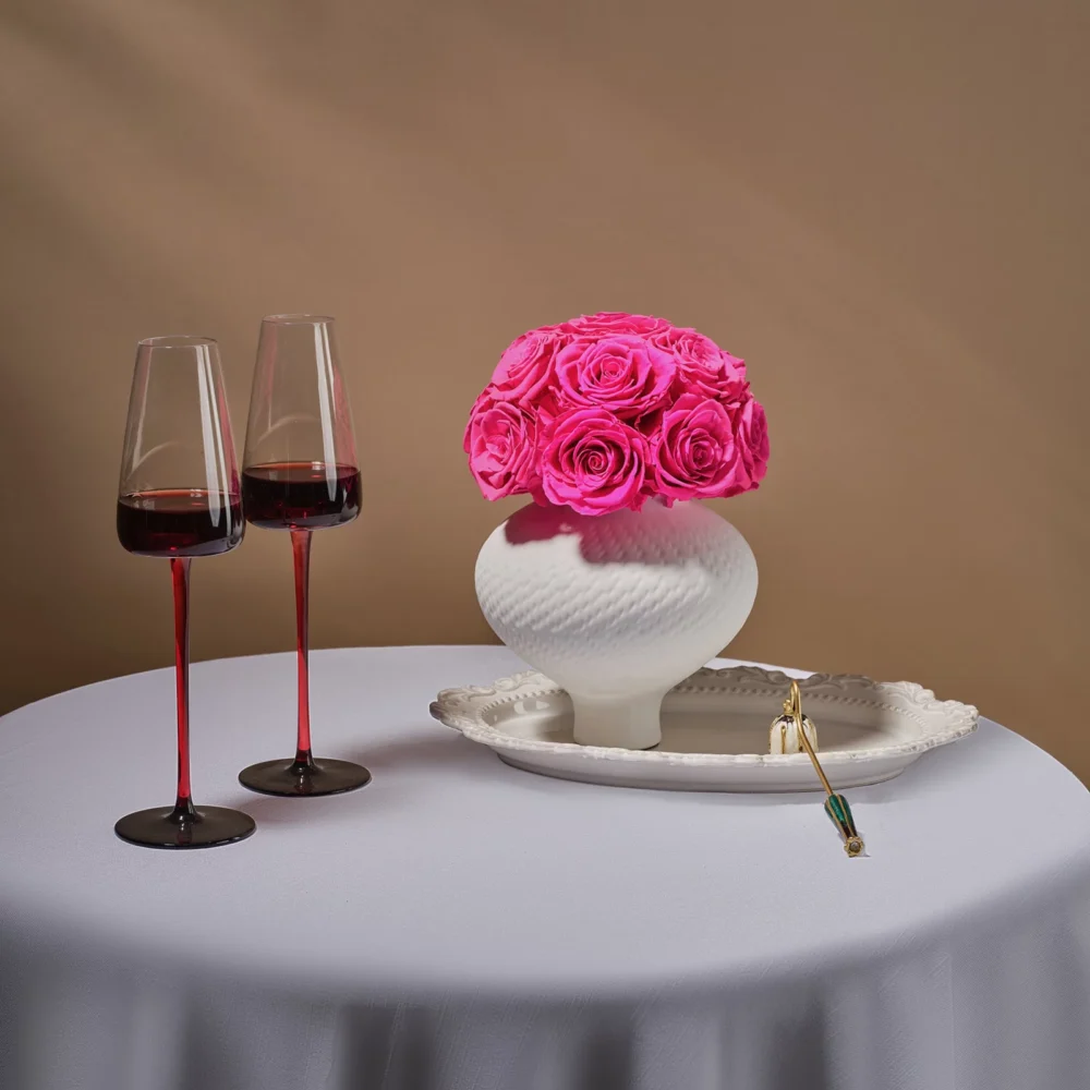 White ceramic vase with pink roses beside ornate plate with gold spoon and tulip wine glasses