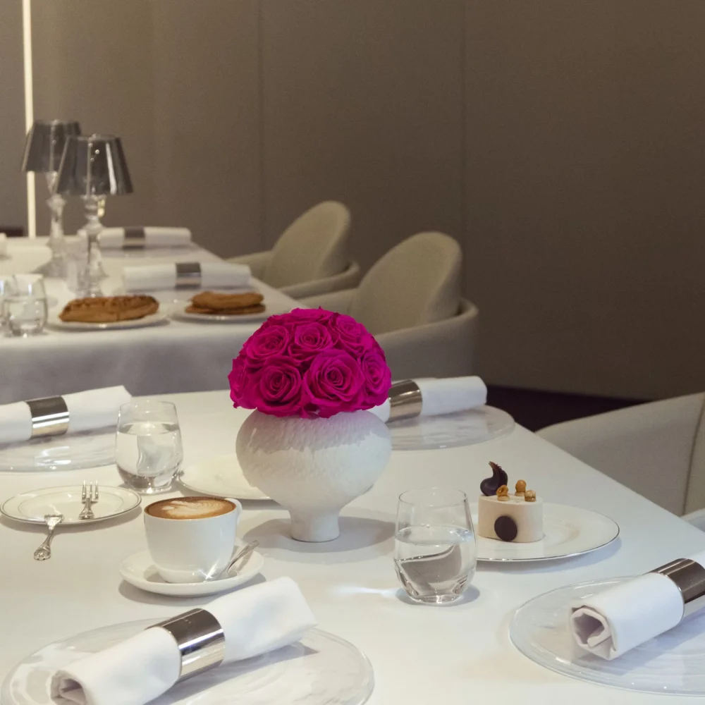This photograph showcases an elegantly set dining table in a modern, minimalist restaurant. The table is draped in a white tablecloth, adorned with a vibrant, pink rose in a white ceramic vase.