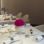 This photograph showcases an elegantly set dining table in a modern, minimalist restaurant. The table is draped in a white tablecloth, adorned with a vibrant, pink rose in a white ceramic vase.