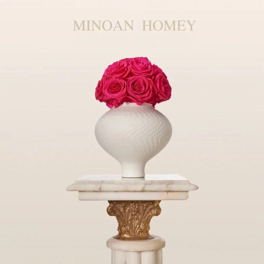 A minimalist white fluted vase with pink roses on a gold-accented marble pedestal, against a cream background. Elegant 'MINOAN HOMEY' text above. "