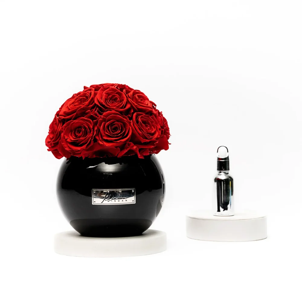 Glossy black vase with silver label holding lush red rose bouquet beside silver perfume bottle on white pedestals